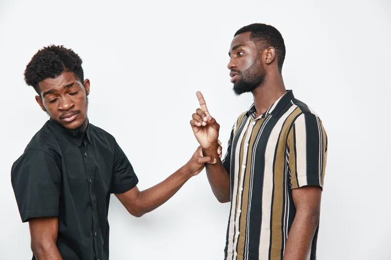 Two African American men with dark skin having a serious conversation, one gesturing with finger, casual clothing on white background in studio.