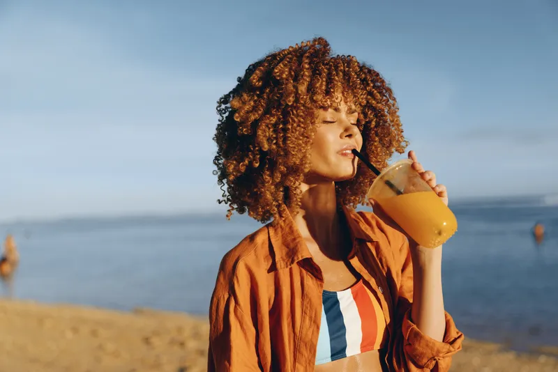 A young woman with curly hair enjoys a fresh orange juice on the beach under clear blue sky. She wears a colorful swimsuit and a light orange shirt, relaxing by the sea.