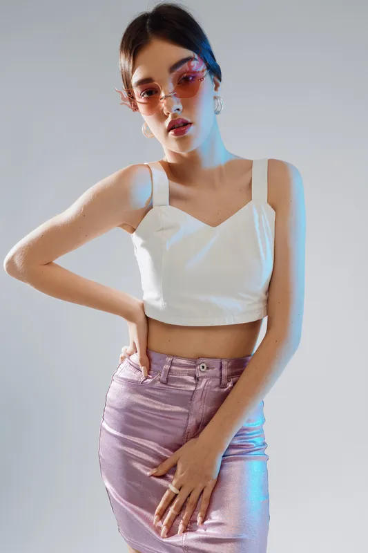 Young woman in a white crop top and shiny pink skirt poses confidently wearing pink sunglasses. Studio shot with soft lighting. Concept of woman beauty fashion and style.