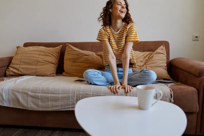 A woman sits crosslegged on a brown couch in a cozy living room, smiling and relaxed. A warm, casual home lifestyle moment captured in natural light, highlighting comfort, everyday routine, and
