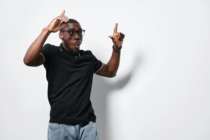 African American man with dark skin wearing glasses and black polo shirt posing with hands up and surprised expression on white background. Casual style and modern look.