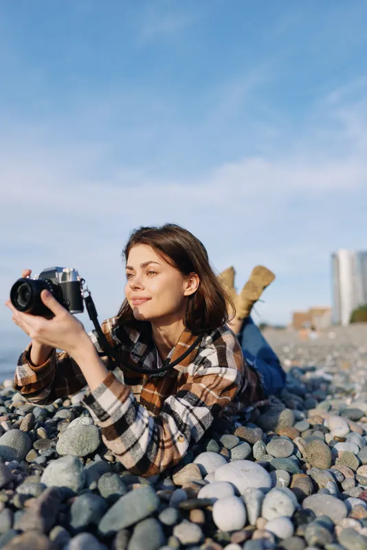 Female photographer lying on a rocky beach, examining a camera, plaid shirt, sunny day, distant skyline, focus on creativity and outdoor storytelling.