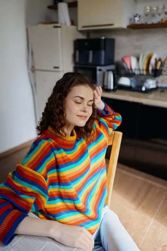 Young woman with curly hair relaxes in a bright kitchen, wearing a colorful striped sweater and resting her head on her hand, creating a calm, cozy domestic moment.