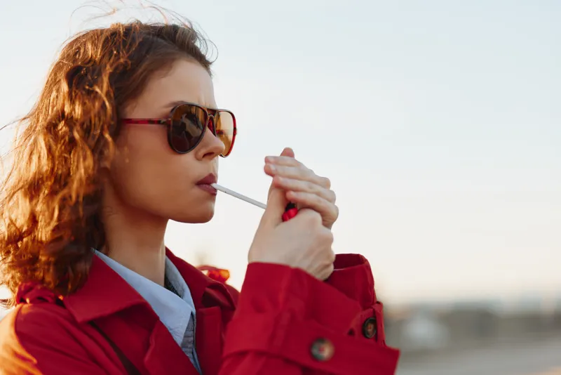 A young woman in a red coat and sunglasses lights a cigarette outdoors during daylight, focusing on the flame and cigarette. Concept of smoking habit and urban lifestyle.