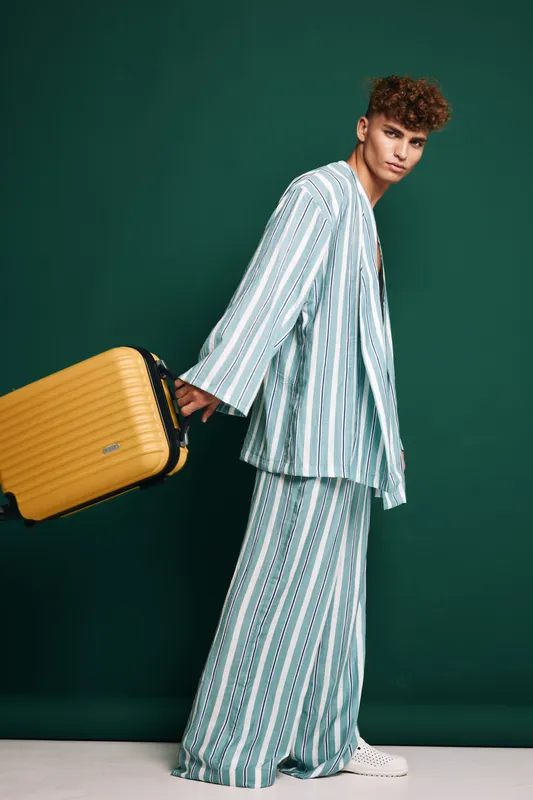 Young man in striped pajamas stands with rolling yellow suitcase against green background holding handle with casual confident expression and curly hair style in studio.