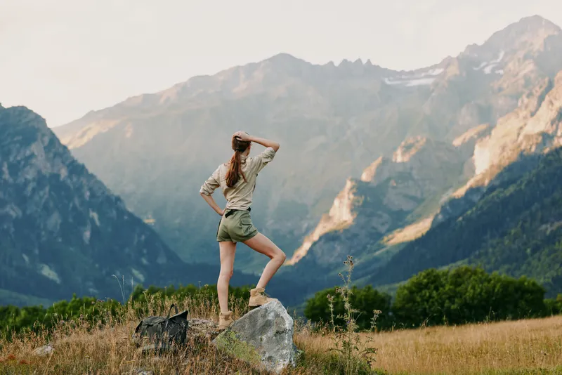 A woman stands on a rock in an alpine meadow, looking toward distant peaks, capturing a moment of outdoor adventure, personal exploration, and calm freedom.