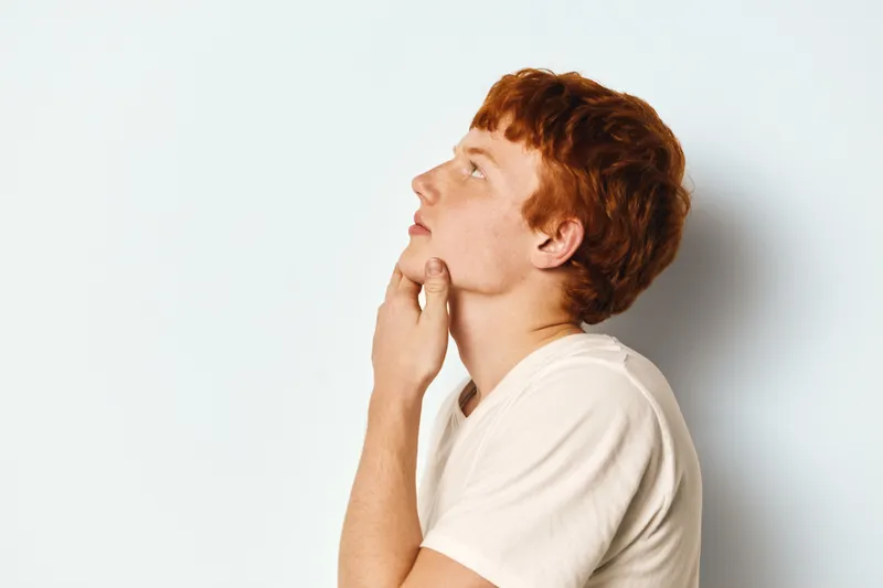 Young man with ginger hair and fair skin wearing casual t-shirt looking thoughtful on white background, side view of profile for contemplation and idea concept.