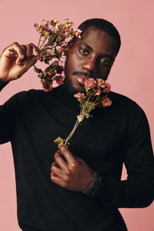Black man with dark skin holding dried flowers in creative conceptual visual style on pink background. Portrait of African ethnicity male with artistic expression and fashion black turtleneck.