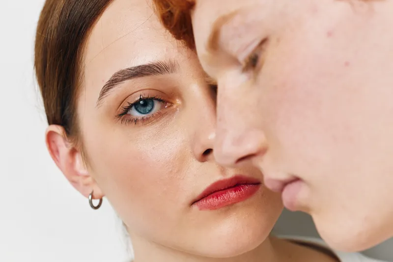 Closeup of two young people with clear skin and focused expression, natural light, minimal makeup, intimate portrait with shallow depth of field on white background.