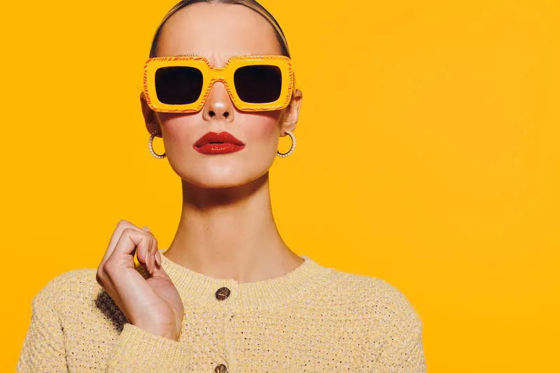 Stylish woman with slicked back hair wears yellow sunglasses and beige sweater against bright yellow background. Fashion portrait with confident expression and bold makeup. Modern style concept.