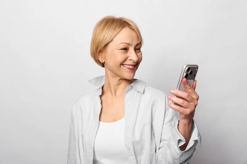 Smiling senior woman holding a smartphone, casual studio portrait on a white background, relaxed communication and technology concept, soft even lighting