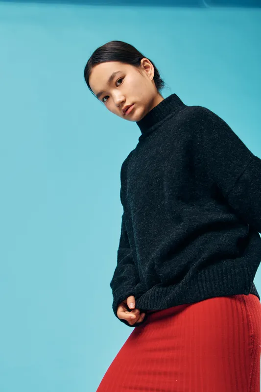 Young woman in a black sweater and red skirt posing against blue background, looking serious with minimal makeup and sleek hair pulled back in a studio portrait.