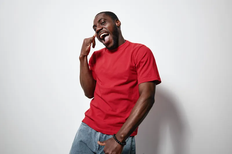 African American man with dark skin wearing red t shirt and jeans posing happily with hand gesture and smiling against white background in studio. Casual style and joyful expression.