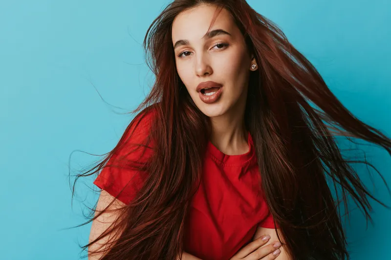 Young woman with long brown hair wearing red shirt posing with flowing hair and expressive face on blue background. Beauty, fashion and portrait with casual style and natural look.