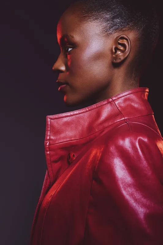 Profile portrait of confident black woman with dark skin wearing red leather jacket against dark background. Concept for fashion, style, and beauty with dramatic lighting and modern look.