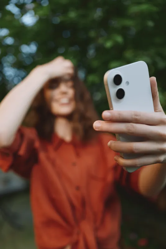Young woman with curly hair takes a selfie outdoors among lush trees, capturing a bright, candid moment and a relaxed, confident mood