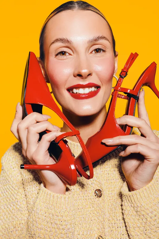 Smiling woman with red lipstick holds red high heel shoes near face on bright yellow background. Concept of fashion, style, beauty, and vibrant color contrast.