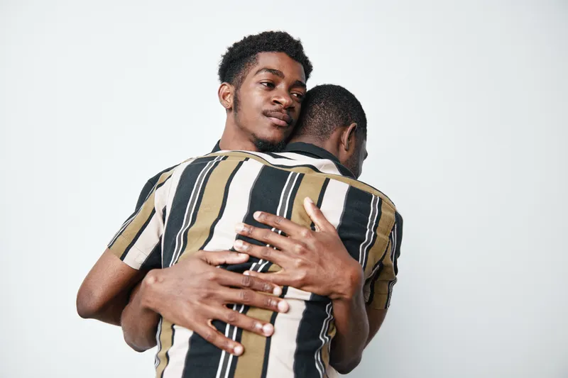Two young black men with dark skin hugging each other in emotional embrace on white background. Male friends showing support, connection, and affection with casual striped shirts.