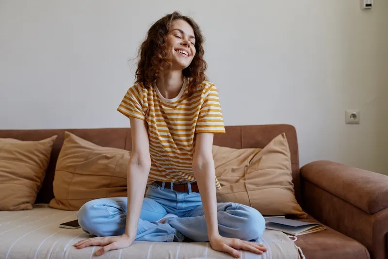 A relaxed woman at home sits on a sofa in a striped yellow t shirt and denim jeans, smiling softly. A candid lifestyle moment celebrating comfort and everyday happiness.