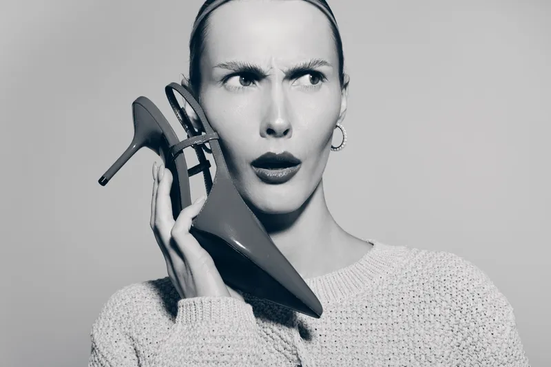 A white woman with pale skin and dark eyebrows holds a high heel shoe to her ear with a confused expression. Concept for fashion, communication, and unusual objects in studio.