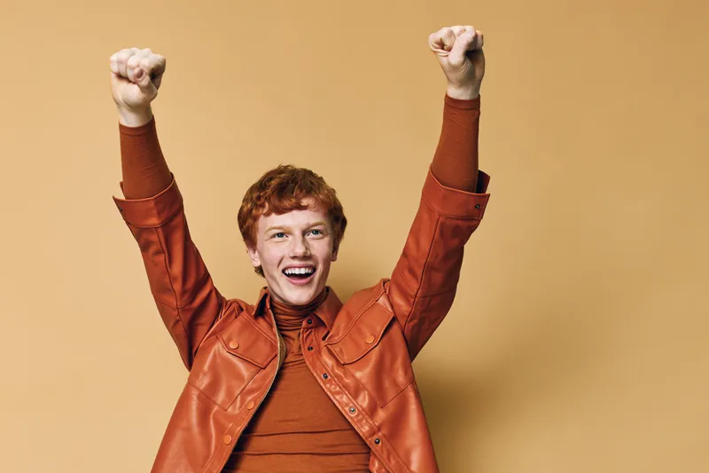 Happy young man with red hair celebrating success with raised fists wearing a brown jacket and sweater on beige background with joyful expression and excitement.