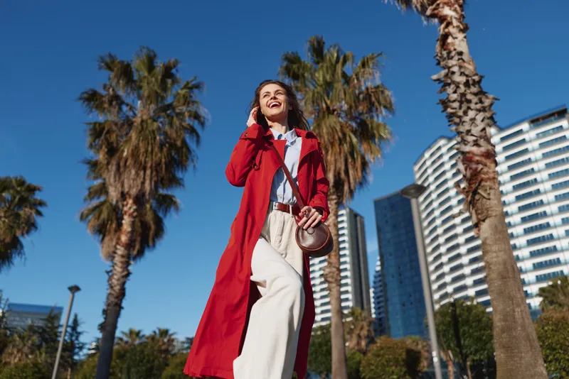 Young woman in a red coat walks through a sunny urban street, smiling confidently as palm trees and modern buildings loom in the bright background.