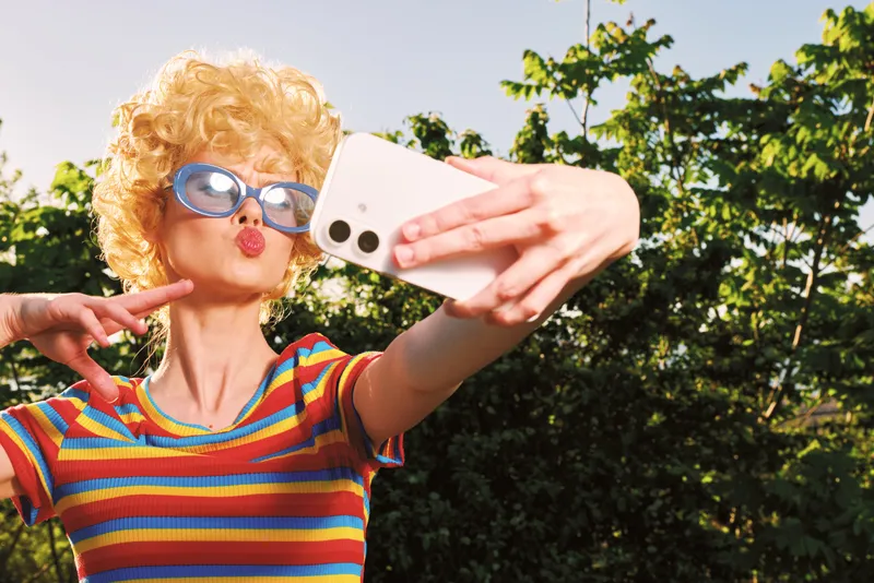 Selfie of a woman with curly hair wearing a striped shirt, blue sunglasses, outdoors on a sunny day. Retro film color, playful mood, self expression, casual lifestyle.
