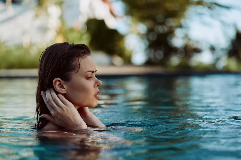 Relaxed woman in pool, shoulders above water, thoughtful expression, outdoor lifestyle scene, soft sunlight, summer leisure, calm moment, reflective mood, serene backdrop and calm setting.
