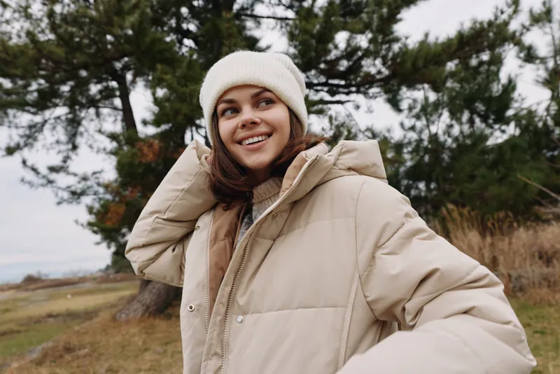 Woman outdoors in a beige puffer jacket and knit beanie, smiling in a park with trees and open field, depicting casual winter style for lifestyle and fashion photography.