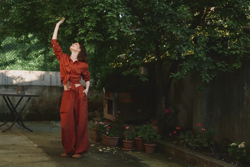 Joyful woman outdoors in a red jumpsuit, raising her hand toward the sun, relaxed posture in a lush garden, soft light, natural colors, candid lifestyle moment