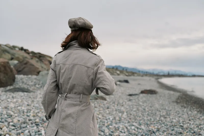 Woman in checkered coat and beret walks along rocky beach under cloudy sky. Autumn or winter outdoor scene with calm sea and distant mountains in the background. Casual fashion style.