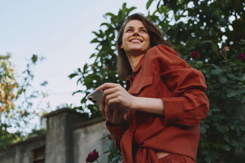Cheerful woman outdoors, lifestyle moment with a smile, red clothing, phone in hand, garden setting, casual pose, sunlit mood, natural colors, candid portrait, fresh breeze