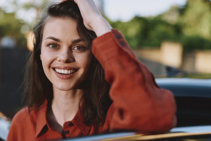 Smiling woman in a casual orange shirt outdoors by a car, captured in a candid lifestyle moment with natural daylight and joyful expression that conveys warmth