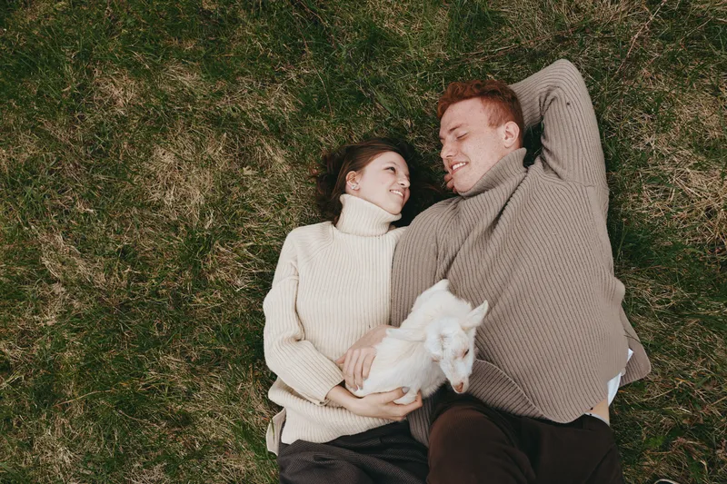 A warm couple lies on green grass, cuddling a white goat kid, wearing knit sweaters. An outdoor lifestyle moment of bonding, joy, and affection in a pastoral setting.