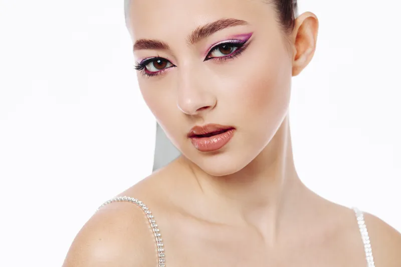 Close-up of a young woman with bold colorful makeup and glossy lips. Clean white background highlights artistic beauty and fashion creativity in studio lighting.