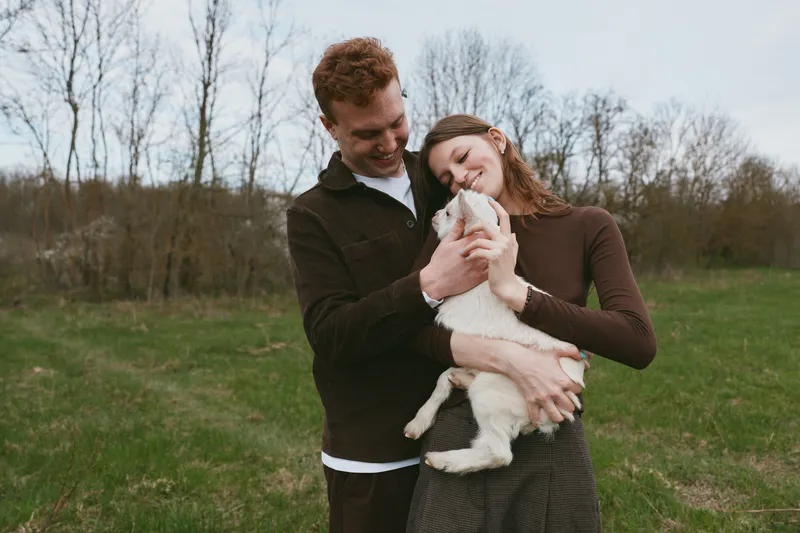 A young couple embraces a white baby goat outdoors in a grassy field during early spring, sharing a tender moment of connection and joy in a casual lifestyle setting.