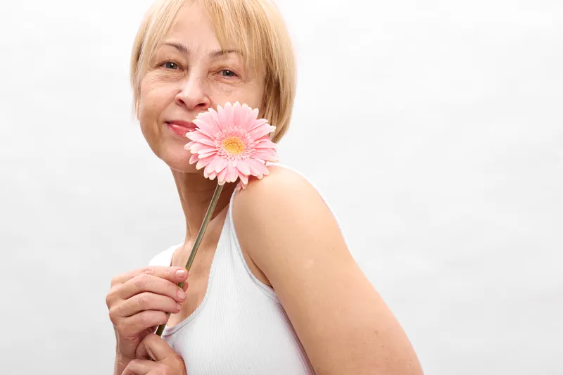 Woman holding a pink flower blossom in close up portrait, blonde hair, spring mood, studio white background, soft smile, gentle light, minimal scene calm