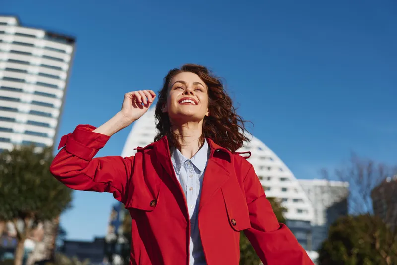 Young woman with curly hair enjoys sunny weather outdoors wearing a red coat and smiling against a clear blue sky in an urban environment.