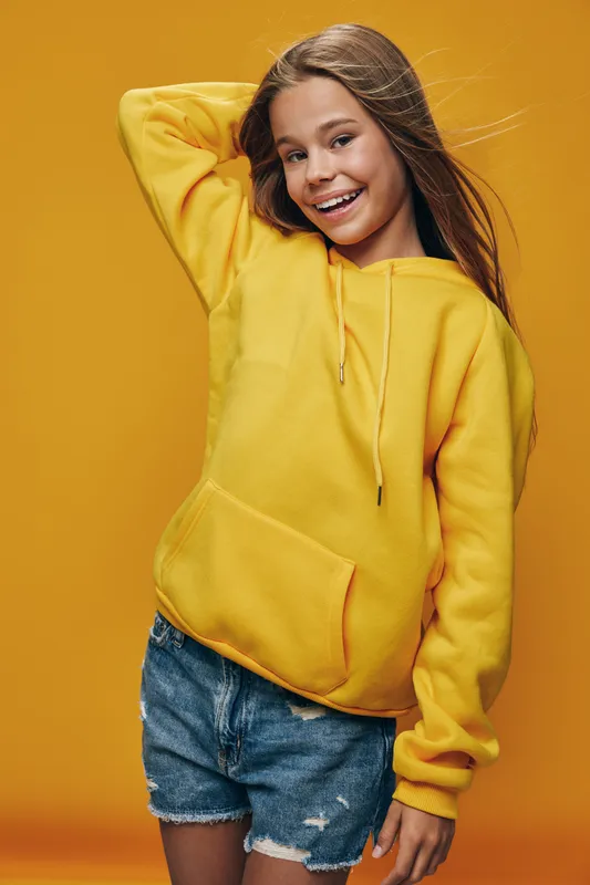 Girl smiling and posing in yellow hoodie with denim shorts on orange background. Long hair, casual style, cheerful young female with one hand behind head in studio.