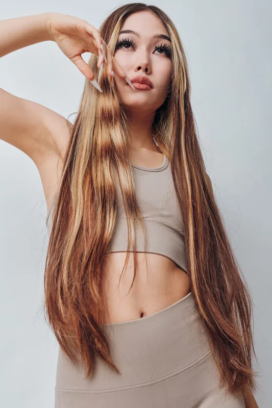 Woman with long straight hair posing in casual sportswear top and leggings, looking upwards with hand near face against plain background. Fitness, beauty, casual and fashion portrait.