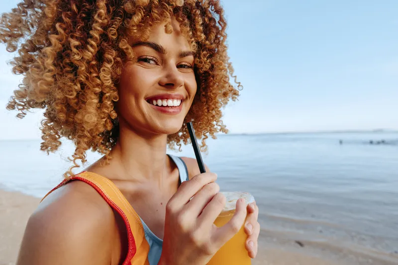 A smiling woman with curly hair on the beach sips a bright drink through a straw, enjoying a sunny summer moment. A relaxed lifestyle scene focused on leisure.