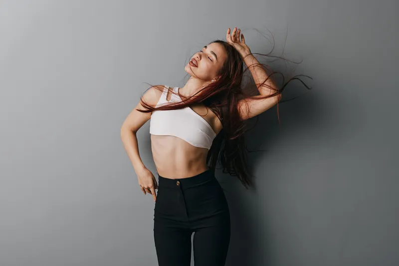 Young woman dancing with closed eyes and long hair flowing in motion against gray background. Casual outfit with white crop top and black pants showing active lifestyle and joy.