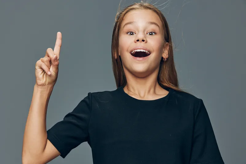Girl smiling and pointing upward with finger, looking excited and surprised, wearing black shirt against gray background with positive expression.