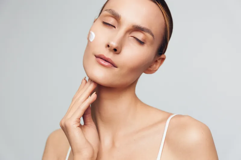 Young woman with closed eyes applying face cream, serene expression, smooth skin, natural beauty, minimal makeup, soft lighting, clean background, skincare routine