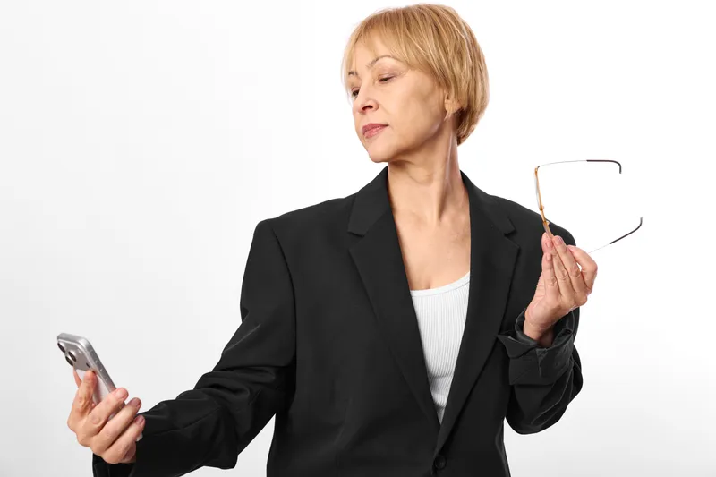 Woman in business suit holding glasses and smartphone, studio on white background, professional communication, concerned expression, concept of focus, vision, stress, modern work life, soft light