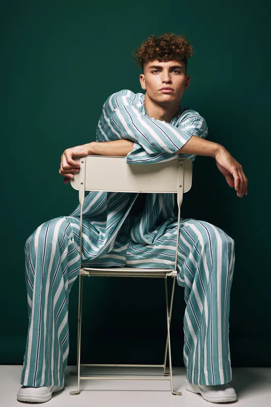 Young man with curly hair wearing striped casual outfit sitting backwards on chair against green background, posing with confident expression and relaxed posture.