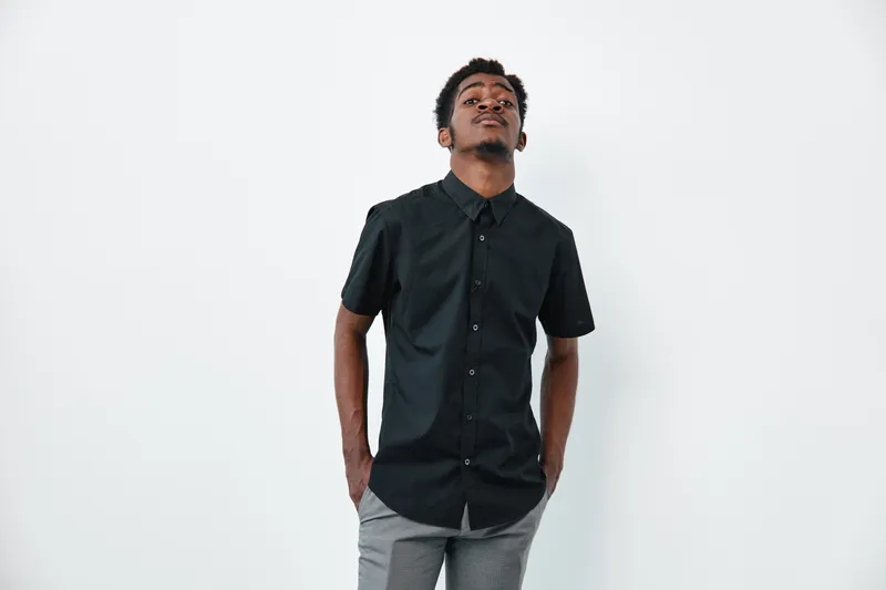 Young black man with dark skin wearing black shirt and gray pants standing with hands in pockets against white background looking confident.