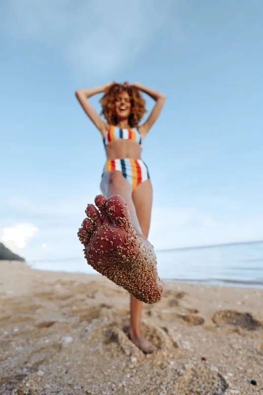 Curly haired woman in striped swimsuit poses on beach, arms on head, sand and mud on legs, summer outdoors fitness concept, ocean breeze and horizon in background