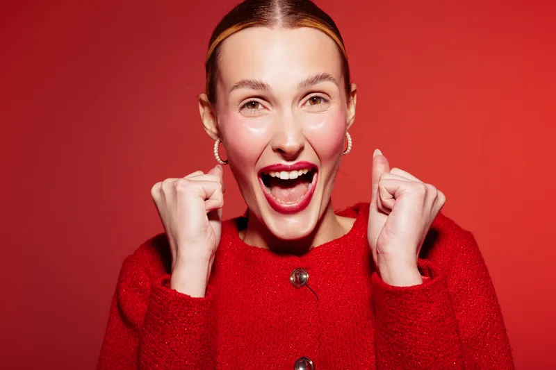 Excited woman in red sweater smiles widely with clenched fists on red background. Concept of happiness, celebration, success, and positive emotions in studio lighting.