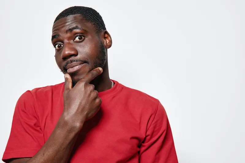 African American man with dark skin wearing red shirt looking thoughtful with hand on chin isolated on white background. Portrait of pensive black male with short hair and beard.